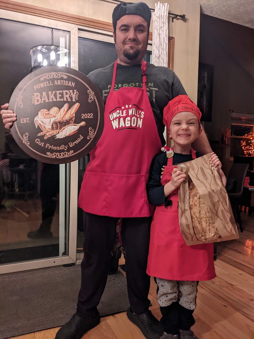 Allen holding the Powell Artisan Bakery sign beside his son, who is wearing a chef's hat and holding a bagged loaf