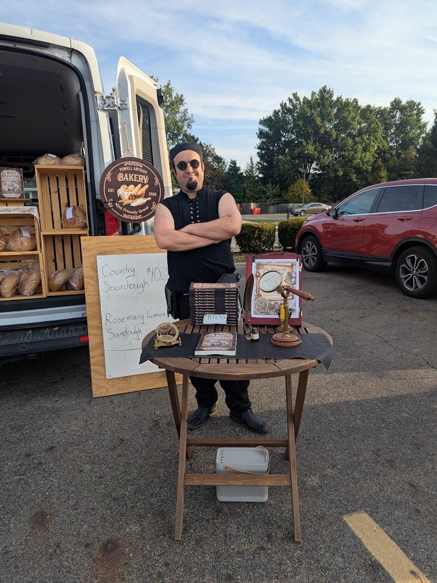 Allen at the market stall behind a table holding both loaves of bread and stacked copies of his book The Price of Discovery