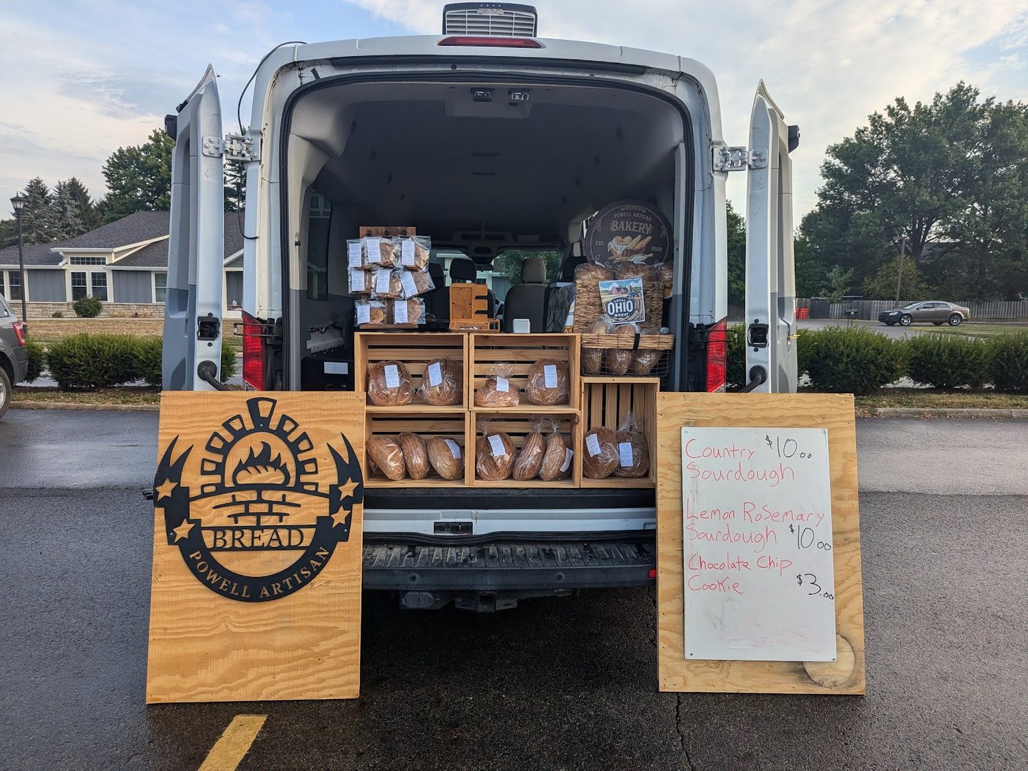 The back of a cargo van set up as a market stall, crates of bagged sourdough, a hand-lettered price board, and a Powell Artisan Bakery sign